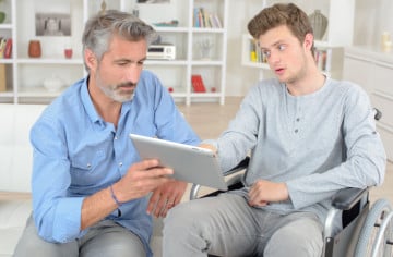 Older man supporting younger man in wheelchair who is using a tablet